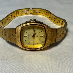 Peut inclure: Une montre Citizen Quartz dorée avec une finition texturée sur le boîtier et le bracelet à maillons. La montre présente un cadran carré aux coins arrondis, un fond doré, des index noirs fins et des aiguilles noires. Le texte Citizen Quartz est visible.