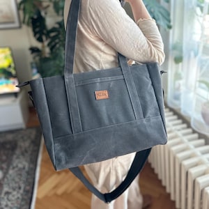 Może przedstawiać: Szara, płócienna torba typu tote z brązową skórzaną naszywką przedstawiającą rybę. Torba ma długi, regulowany pasek i górne zapięcie na zamek błyskawiczny.