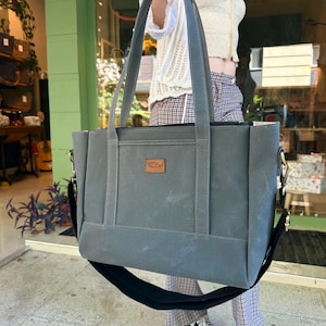 May include: A gray canvas tote bag with black straps and a brown leather patch. The bag has a zippered closure and a top handle.