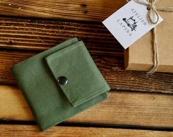 May include: An olive green canvas wallet with a snap closure sits on a wooden surface. A small gift box tied with twine and a tag with the "Atelier Lapina" logo are also present. The wallet is a compact, square design.