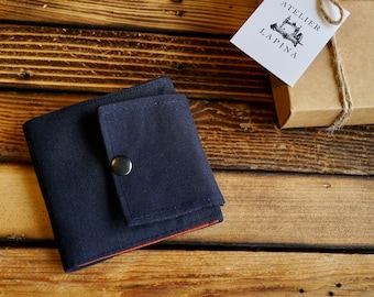 May include: A dark blue fabric wallet with a small pocket on the front, secured with a button. The wallet has a red lining and is sitting on a wooden surface. The text "Atelier Lapina" is visible on a brown gift box in the background.