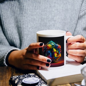 May include: A white ceramic mug with a black background and a colorful abstract design of a cube. The mug is being held by a person's hands.