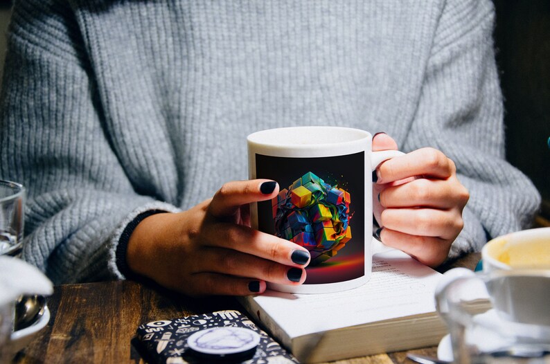 May include: A white ceramic mug with a black background and a colorful abstract design of a cube. The mug is being held by a person's hands.
