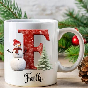 May include: White ceramic mug with a red and white snowman, a green pine tree, and a red letter F with white snowflakes. The mug has the name "Faith" written in black at the bottom.