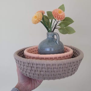 Może przedstawiać: Zestaw trzech plecionych koszy w różnych rozmiarach i kolorach. Największy kosz jest jasnobrązowy, środkowy różowy, a najmniejszy zawiera niebieski wazon z pomarańczowymi i żółtymi kwiatami.