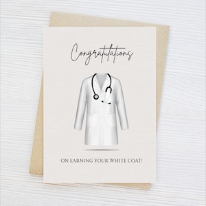 May include: A greeting card with the word "Congratulations" in elegant script. The card features a white lab coat and stethoscope illustration, with the text "ON EARNING YOUR WHITE COAT!" below. The card is on a beige envelope.