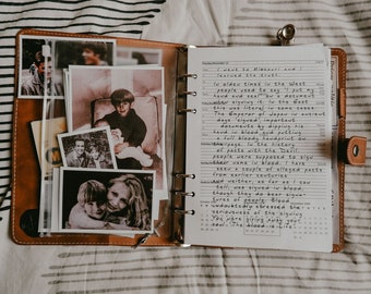 May include: Open brown leather journal with handwritten notes and several black and white photographs. The journal is open to a page with text and a calendar. The photos show people.