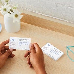 Customizable allergy alert cards.  Includes, name, allergen, restaurant instructions, table number.  Showing both sides with one card in a persons hands.  Other cards on a desk with eye glasses and blue clips.