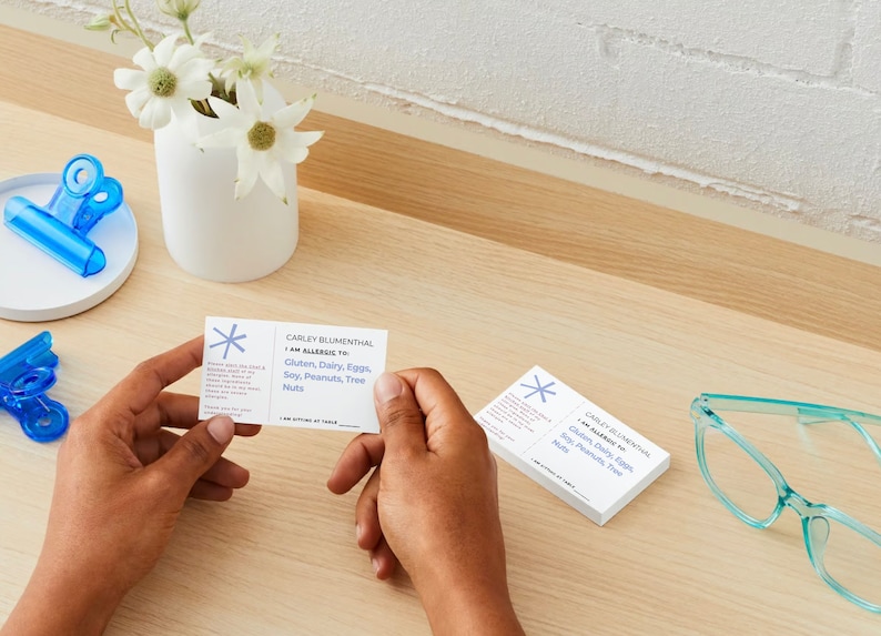 Customizable allergy alert cards.  Includes, name, allergen, restaurant instructions, table number.  Showing both sides with one card in a persons hands.  Other cards on a desk with eye glasses and blue clips.