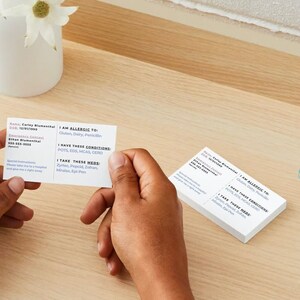 Customizable medical alert cards.  Includes, name, allergens, EMS instructions, conditions, medications, emergency contact, DOB.  Shows both sides of card.  A person is holding a card and the others are on a wood desk.