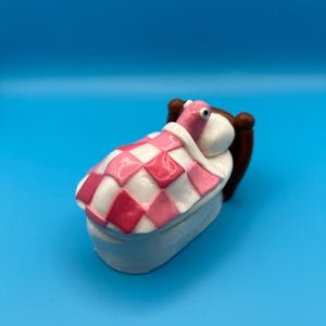 May include: A miniature bed with a sleeping creature. The bed frame is white, and the bedding has a pink and white checkered pattern. The creature is pink with a white head and a brown headboard.