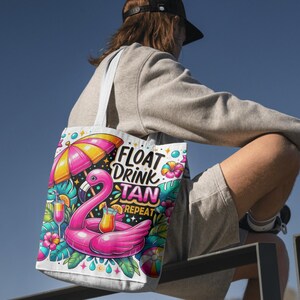 Könnte beinhalten: Weiße Tragetasche mit einem farbenfrohen Design eines pinkfarbenen Flamingo-Schwimmers, einem rosa-gelben Sonnenschirm und den Worten "FLOAT DRINK TAN REPEAT". Tropische Blumen, Getränke und ein Strandball sind ebenfalls abgebildet. Die Tasche hat weiße Träger.