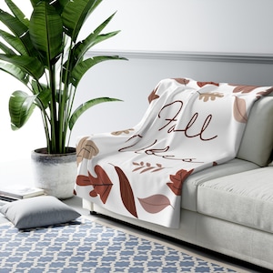 Puede incluir: Manta blanca con temática otoñal, con hojas marrones y beige y la frase "Fall Vibes" en cursiva. La manta está sobre un sofá blanco, con una gran planta verde al fondo.