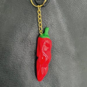 May include: A red chilli pepper keyring with a green stem and gold chain. The pepper is glossy and appears to be made of resin. The background is a textured black surface.