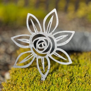 May include: A metal garden stake with a cut-out design of a stylized flower. The flower has a circular center and six petals. The stake is placed in a bed of green moss.