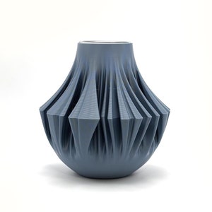 Könnte beinhalten: Eine blaue Keramikvase mit einem gefalteten, geometrischen Design. Die Vase hat einen breiten Boden und eine schmale Öffnung.