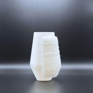 May include: White ceramic vase with a textured surface. A white, textured fabric is draped over the side of the vase.