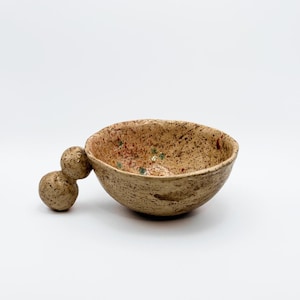 Peut inclure: Un petit bol en céramique beige moucheté avec une poignée arrondie unique. Le bol est recouvert d'un glaçage brun moucheté avec des touches de rouge et de vert.