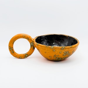 Peut inclure: Un bol en céramique fait à la main avec un design unique. Le bol est orange avec un intérieur noir et une poignée en forme d'anneau. L'extérieur présente des mouchetures vertes. C'est un objet décoratif.