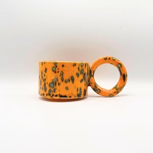 May include: Orange ceramic mug with a unique circular handle. The mug features a speckled pattern of dark green and black spots across its surface. The mug is designed with a cylindrical body and a rounded handle, perfect for enjoying hot beverages.