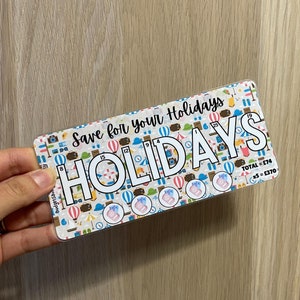 May include: A clear plastic savings tracker with a colourful design featuring travel icons. The tracker has a section for saving money for holidays with the text "Save for your Holidays" and "HOLIDAYS". The tracker has a total of 19 spaces for saving money, with a total of £74 and a *5 = £370.