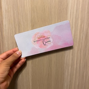May include: A rectangular card with a watercolour design in shades of pink and white. The card features the text "BUDGET SAVING Queen" in black script, with a floral illustration. The card is held against a wooden background.