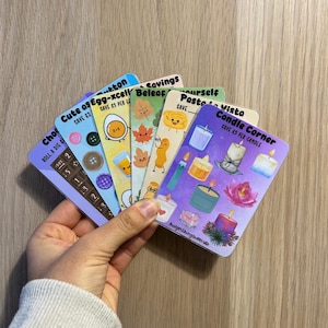 May include: A hand holding a stack of colourful cards with various designs. The cards feature illustrations of candles, food items, and text such as "Candle Corner" and "Cute as Egg-xcell". The cards are on a light brown wooden surface.