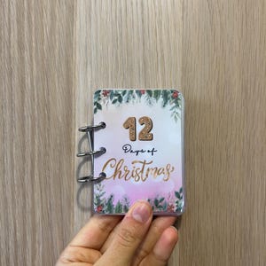 May include: A small, rectangular Christmas booklet with a clear plastic cover. The cover features the text "12 Days of Christmas" in gold script, with a gingerbread-style "12" and festive greenery. The booklet is held open by a silver binder.