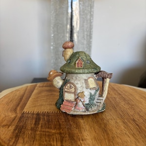 Könnte beinhalten: Ein kleines Keramik-Pilzhaus mit grünem Dach, brauner Tür und einer kleinen Figur eines Menschen, der davor steht.