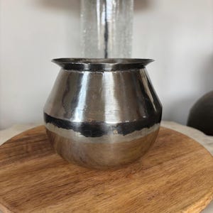 Puede incluir: Una pequeña olla de metal plateado con un cuerpo ancho y redondeado y un borde ligeramente ensanchado. La olla está sobre una tabla de cortar redonda de madera. El metal tiene una textura martillada y refleja la luz.