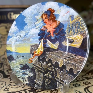 May include: Decorative plate featuring a woman in a blue dress, seated on a stone wall overlooking the sea. The artwork includes a castle, a flowing yellow scarf, and a blue sky with clouds. The plate is supported by a clear plastic stand.