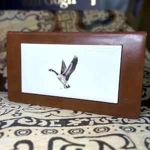 Puede incluir: Un marco de fotos rectangular de cuero marrón con un inserto blanco que presenta una ilustración en blanco y negro de un ganso en vuelo. El marco tiene un diseño simple y clásico, adecuado para exhibir una pequeña fotografía o una obra de arte.