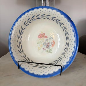Puede incluir: Un tazón de cerámica blanco vintage con un borde azul y un diseño floral. El tazón tiene un fondo blanco con un patrón azul y blanco alrededor del borde y un diseño floral en el centro.