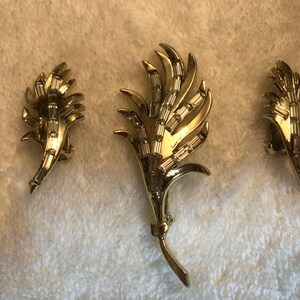 Op de afbeelding: Een set van drie gouden sieraden met een gestileerd bladontwerp met heldere strass steentjes. De set bevat een broche en een paar oorbellen.