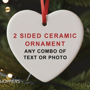 May include: A white heart-shaped ceramic ornament with red text that reads "2 SIDED CERAMIC ORNAMENT ANY COMBO OF TEXT OR PHOTO." A red ribbon is attached for hanging. The ornament is displayed against a backdrop of a Christmas tree.