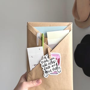 May include: A brown paper organiser with a folded design, holding stationery items. A sticker with the text "At the table, you will never lack any good thing" is attached with a wooden clip. Pens and paper are visible.