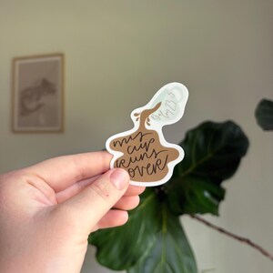 Puede incluir: Una pegatina con la frase "my cup runs over" en una fuente manuscrita. La pegatina tiene un diseño de taza de café marrón con un contorno blanco. La parte superior de la taza tiene un diseño azul claro con el texto "Psalm 23:5".