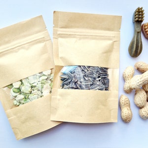 May include: Two brown paper bags with clear plastic windows, one filled with green and white pea-shaped snacks, the other with black sunflower seeds.  A pile of peanuts and two green and brown toothbrush-shaped dog chew toys are on a white surface.
