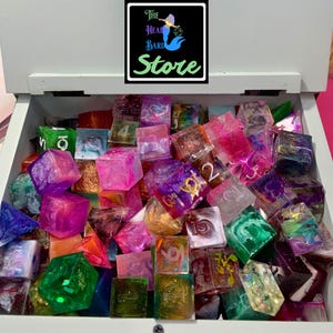 May include: A collection of multi-colored resin polyhedral dice in a white wooden box. The dice feature various swirls, glitter, and translucent finishes with engraved numbers. A black logo with the text The Head Bard Store is visible.