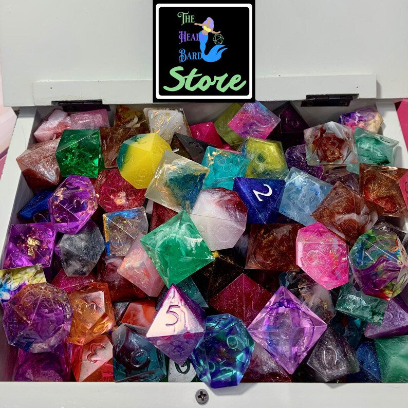 May include: A collection of multi-colored resin polyhedral dice in a white box. The dice feature various swirls, glitter, and metallic inclusions. Each die is engraved with numbers. A black logo at the top reads The Head Bard Store.