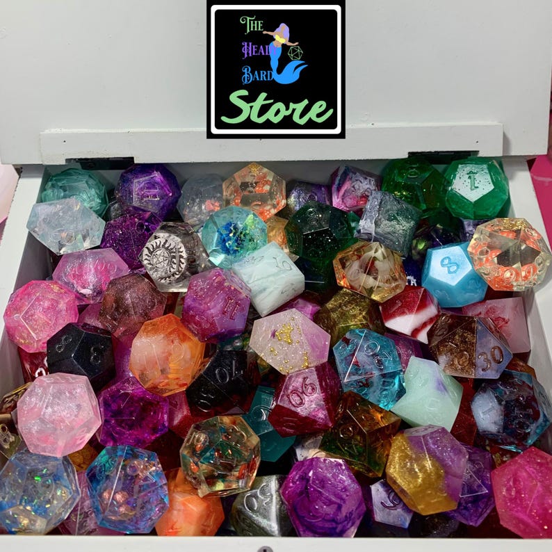 May include: A collection of multi-colored polyhedral dice in a white box. The dice feature various finishes including glitter, translucent, and opaque styles with engraved numbers. A black sign above the box displays the text The Head Bard Store.