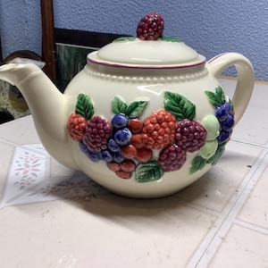 May include: A white ceramic teapot with a lid decorated with a colorful berry design. The teapot has a round body and a spout on the side. The berries are red, purple, and green.