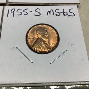 May include: A 1955-S Lincoln penny in a coin holder. The coin is in MS65 condition. The coin is copper colored and has the words "In God We Trust" and "Liberty" on it.