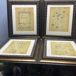 May include: Four framed vintage patent prints. Two prints feature the Monopoly board game layout, while the other two showcase other game designs. The frames are dark brown with gold trim, and the prints have a sepia tone.