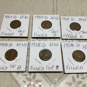May include: Six vintage copper pennies in white cardboard holders. Each holder is labeled with the year, mint mark, and grade. The pennies are from 1957 and 1958. The holders have handwritten notes. The pennies are displayed on a patterned surface.
