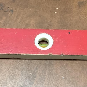 May include: A rectangular red spirit level tool featuring two circular white-rimmed vials containing green liquid. The tool has a white base and a small black rectangular label with text printed on the surface, along with a small hole on the right end.