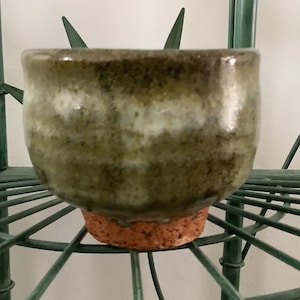 May include: A small, round, green and brown ceramic bowl with a cork base. The bowl has a mottled glaze with white accents.