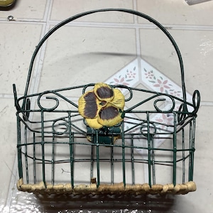 May include: A green metal wire basket with a woven wicker bottom. The basket has a decorative floral design with a yellow and brown flower.