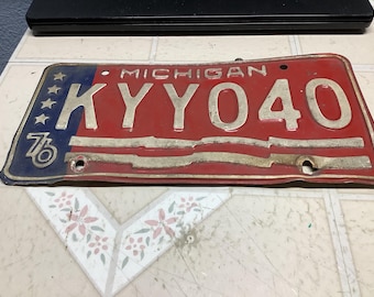 May include: Vintage Michigan license plate with red and blue color scheme. The plate features the state name, the number "76" and the letters "KYY040". The plate has a distressed appearance.