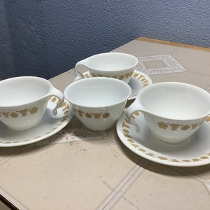 May include: Set of four white ceramic teacups with gold floral designs. Each cup has a matching saucer.